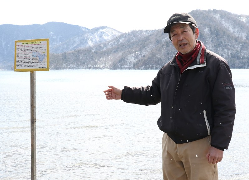 「魚の放射線量をより下げるために検証すべき時期だ」と決意を新たにする中禅寺湖漁協の鹿間久雄専務理事。釣った魚を持ち帰らないように促す看板(左)が立てられている=日光市中宮祠の中禅寺湖で