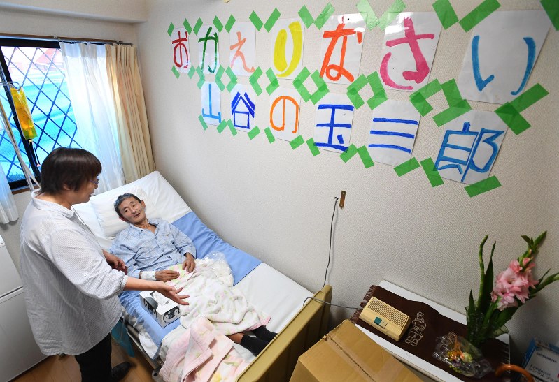 末期の食道がんで入院していた大学病院から山谷のホスピス「きぼうのいえ」へ移り、安心した表情を見せる玉ちゃん。壁には元気だった頃の写真や仲間からのメッセージが飾られていた=中村藍撮影