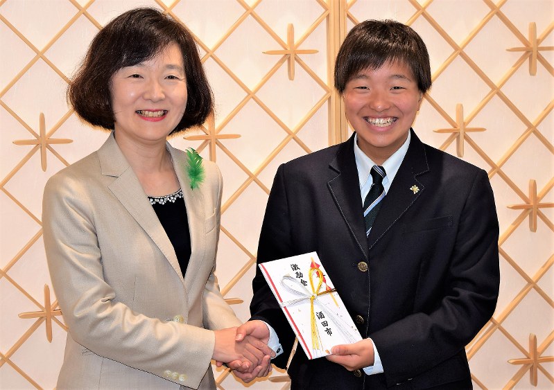 激励金を手に矢口明子副市長(左)と笑顔で握手を交わす斎藤京香さん=酒田市役所で
