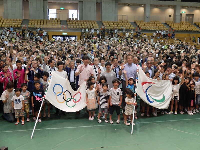 披露された五輪旗とパラリンピック旗を囲む参加者ら=鹿嶋市神向寺の市立カシマスポーツセンターで
