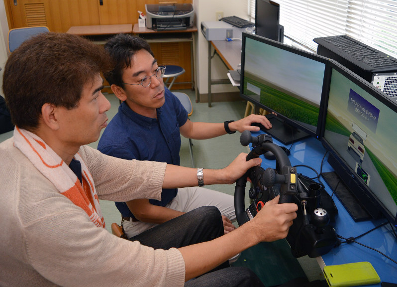 ドライブシミュレーターでの訓練に取り組む浜松市の男性(左)。右はアドバイスする建木健・聖隷クリストファー大助教=浜松市北区の同大で