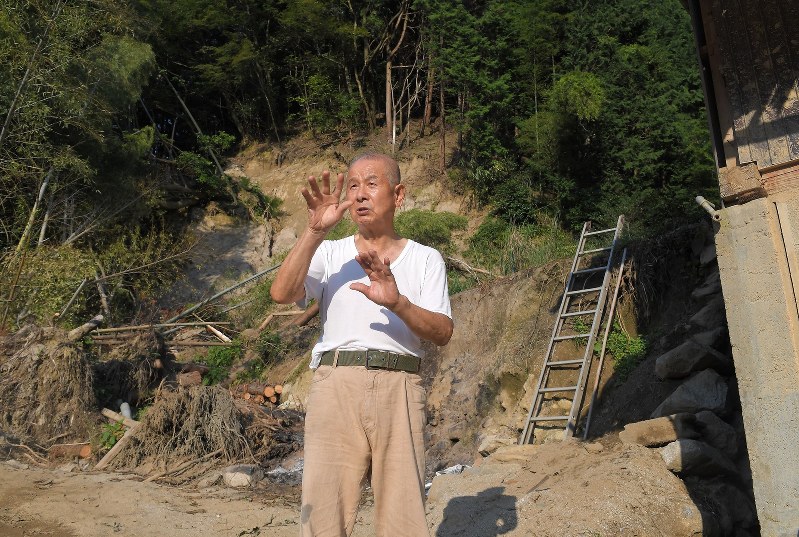 九州北部豪雨後そのままになっている自宅裏の土砂崩れを前に、復興を早く進めてほしいと話す高野博温さん=福岡県朝倉市杷木松末で2017年9月25日、徳野仁子撮影