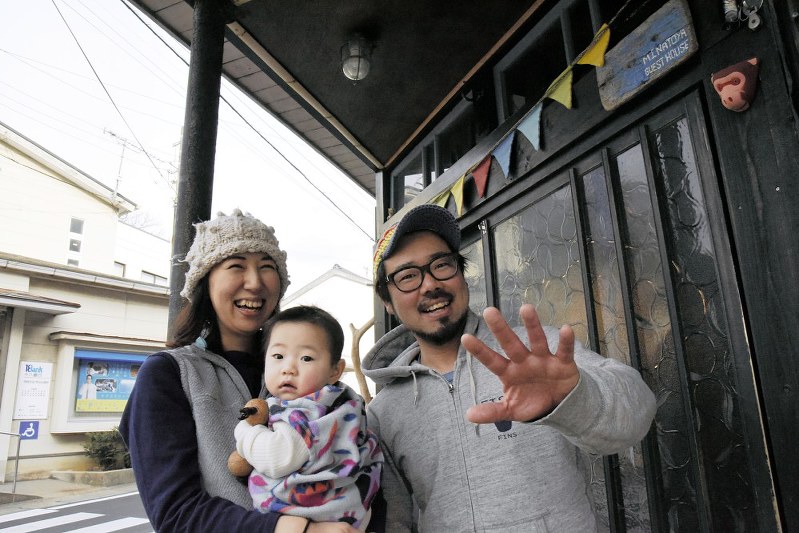 夫婦で開業したゲストハウス「みなとや」の玄関前で、やちよちゃん(中央)を抱いて笑顔を見せる大川香菜さん(左)と漁志さん=長崎県壱岐市で、栗田慎一撮影