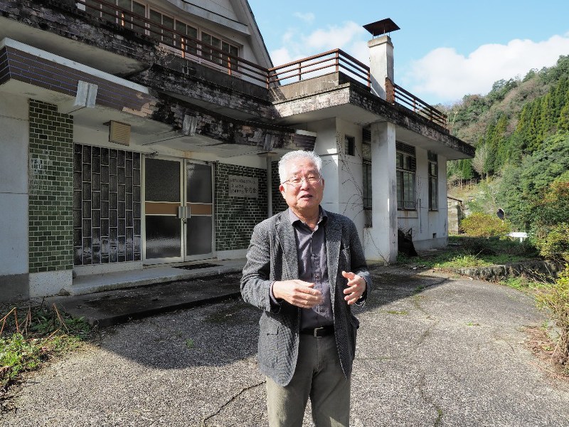 昭和40年代まで使われ、今は放置されている小中学校の寄宿舎の前に立つ山崎一美さん。「市に金がないから、自分たちで頑張れということだ」=島根県益田市匹見町澄川で3月