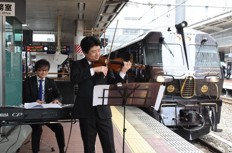 豪華寝台列車の横でテーマ曲を奏でる(左から)光田健一さんと水谷晃さん=JR博多駅で2018年10月16日午前11時21分、宗岡敬介撮影