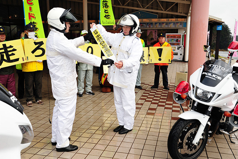 兵庫県警朝来署員から「交通事故ゼロ」の思いが込められたたすきを受け継ぐ京都府警福知山署の山科真琴警部補(右)=京都府福知山市夜久野町の「ドライブインやくの」駐車場で、佐藤孝治撮影