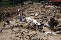 西日本豪雨で消防団の男性が犠牲となった場所に向かって手を合わせた後、手を顔にあてる木坂宮子さん（８２）。近所に住み、草取りなども手伝ってくれたという。「まじめな方だったから一生懸命やってくれたんだと思う」と悼んだ＝広島県呉市安浦地区で２０１８年８月６日午後１時４８分、小川昌宏撮影