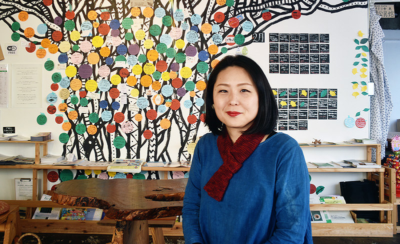 店主の安彦恵里香さん。後ろの壁面には「幸せを呼ぶ木」として人気が高いガジュマルが描かれ、多くの人々のメッセージが所狭しと張られている=広島市中区土橋町2のハチドリ舎で、元田禎撮影