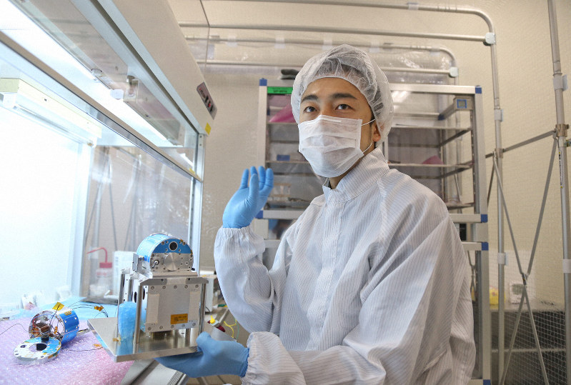 クリーンルーム内で製作中の分離カメラを、レプリカを使って説明する澤田弘崇開発員。左の机上のものは探査機に搭載された本物=相模原市のJAXA相模原キャンパスで2013年、武市公孝撮影