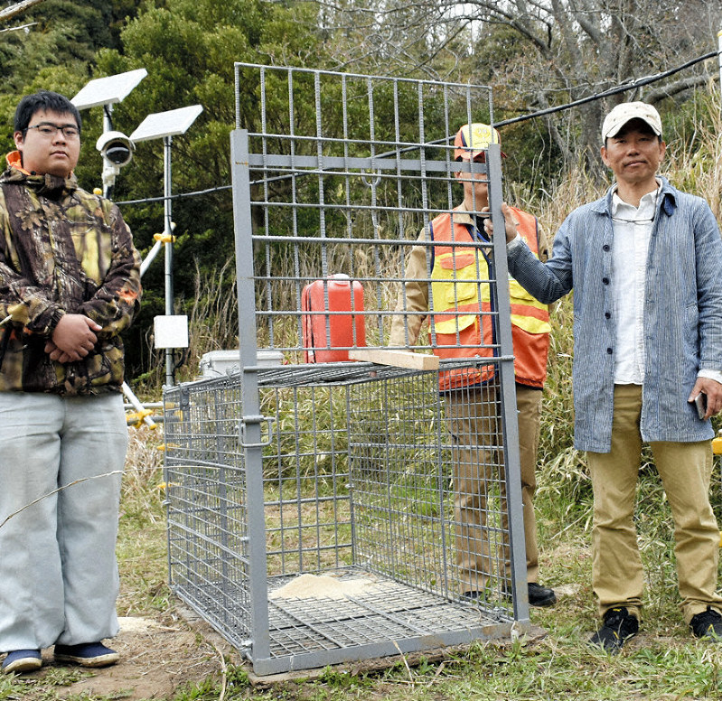 監視カメラなどが設置された捕獲用のおりの横に立つ山野さん(右)ら=木更津市で