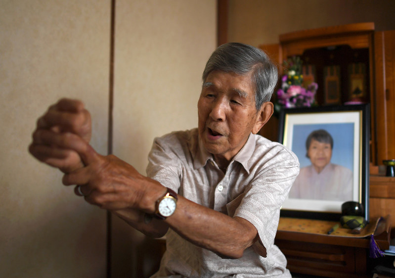 沖縄戦で砲弾の破片が貫通し、今も不自由なままの右腕を示す吉村章さん。右奥は妻晴子さんの遺影=大阪市都島区で2019年6月19日、久保玲撮影