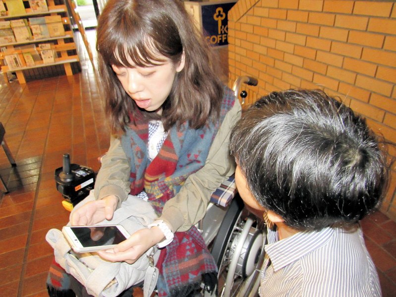 スマートフォンを手に、介助者の女性(手前)と談笑する猪瀬さん=さいたま市中央区で2019年5月7日、夫彰子撮影
