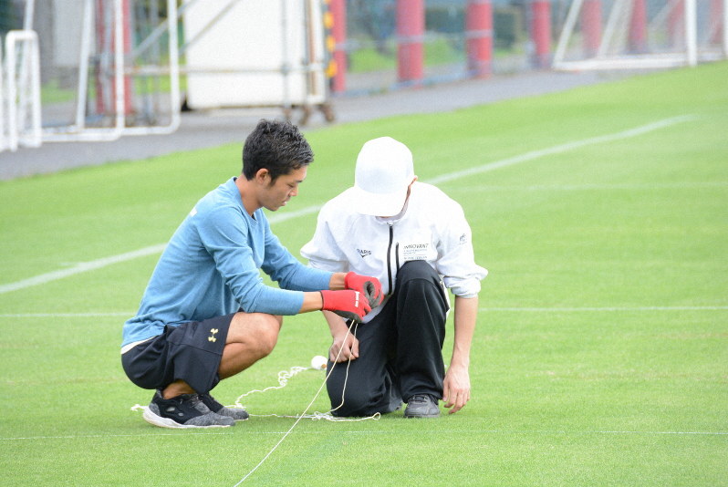 グラウンドキーパー(左)と一緒に、グラウンドに真っすぐラインを引くためのロープを張る少年=東京都小平市大沼町3のFC東京小平グラウンドで2019年7月11日、黒川優撮影