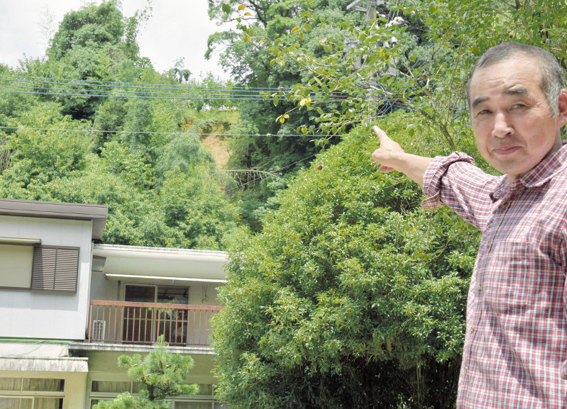 自宅前に立つ小江達夫さん。裏手の斜面は豪雨時に崩れ落ちたままで復旧工事は未着手だ=福岡県朝倉市で、青木絵美撮影