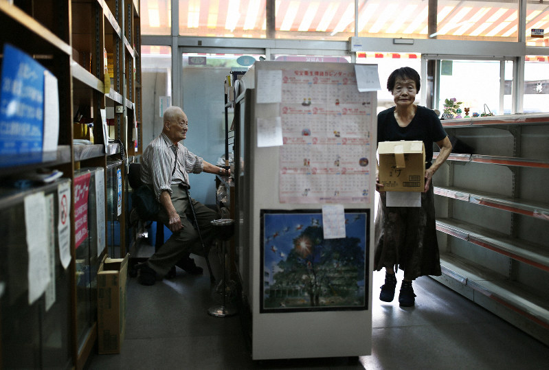 商品が少なくなった店内でレジカウンターの椅子に座る植田義昭さん(左)と片付けをする妻好子さん=大阪府八尾市で2019年9月27日午後4時24分、小出洋平撮影