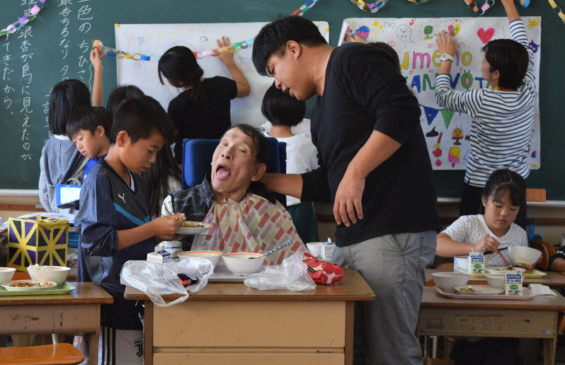 児童に給食を食べさせてもらう田中さん(中央)。後ろは黒板に歓迎の飾り付けをする児童ら。