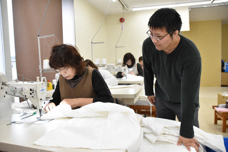 ミシンで膝当ての部分を縫い合わせる従業員の様子を眺める西村圭司工場長(右)=秋田県大仙市で
