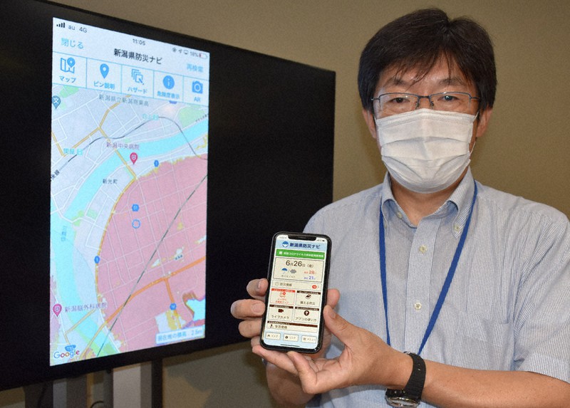 リニューアルしたアプリ「新潟県防災ナビ」を見せる深田課長。背後の画面のように、洪水の浸水想定区域を確認することもできる=新潟市中央区の県庁で