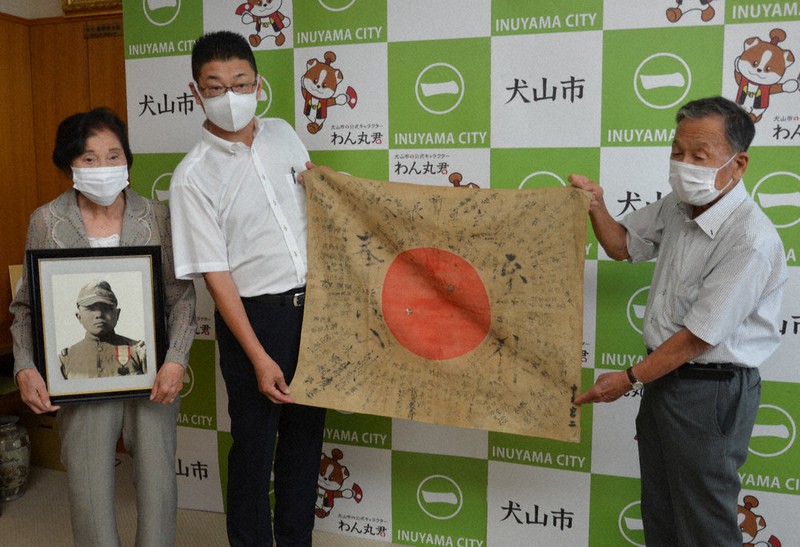 県遺族連合会の柴田義継会長から日章旗を受け取った和泉敬也さん(中央)と則男さんの遺影を持つ芳子さん=愛知県犬山市役所で