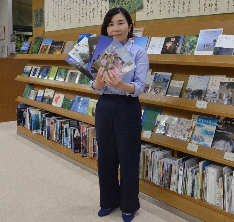 八日市図書館2階の休憩スペースで「そこら」のバックナンバーを手にする山梶瑞穂さん。後ろの書棚には全国各地の地域情報冊子が並んでいる=東近江市八日市金屋の八日市図書館で2020年6月2日午後0時20分、伊藤信司撮影