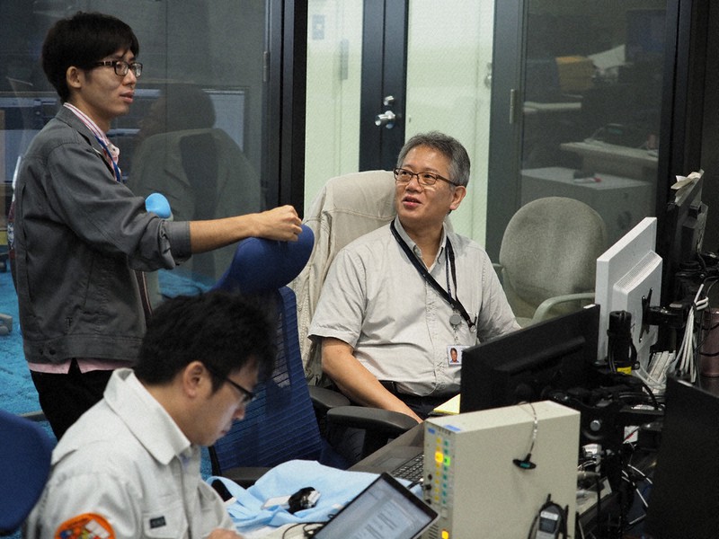 はやぶさ2の運用について、チームの若手メンバーと意見交換するJAXAの照井冬人・特任担当役(右)=JAXA相模原キャンパスの管制室で2018年6月(JAXA提供)