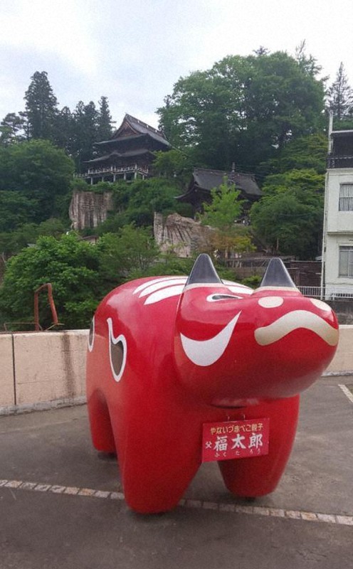 赤べこの発祥地とされる福島県柳津町の公園に置かれた大きな赤べこ。後ろの崖の上に円蔵寺が見える=柳津観光協会提供