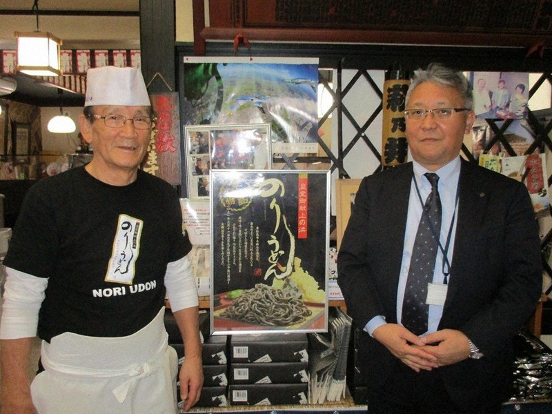 「のりうどん」のポスターの前に立つ「ちゃんこ萩乃井」の大森宣勝会長(左)と石巻商工信組矢本支店長の梶谷忠さん=同信組提供