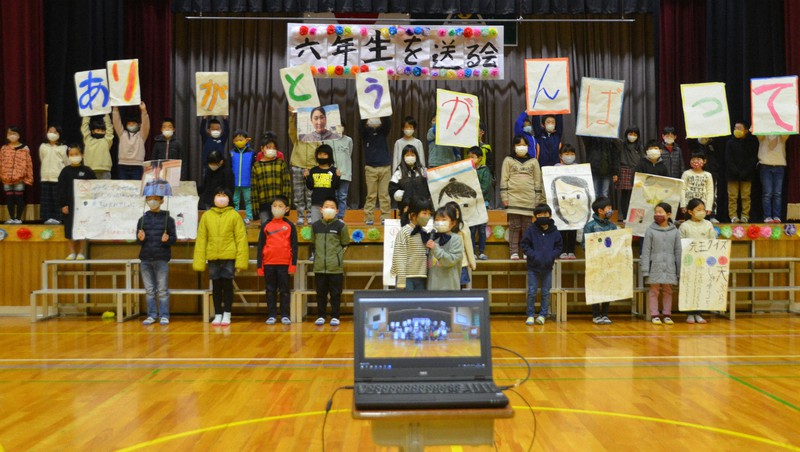 手前に座る6年生に向けて、似顔絵やメッセージを掲げる2年生たち。パソコンなどを使って他学年の教室に生中継された=滋賀県愛荘町島川の秦荘西小で2021年2月24日午前10時14分、伊藤信司撮影