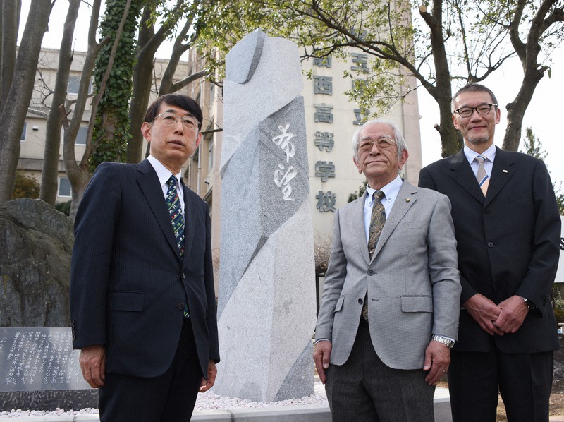 福島県双葉町と埼玉県加須市の友好を記念して建てられた石碑。中央は題字を揮毫した書道家の渡部翠峰さん。右は彫刻家の藤田博史さん、左は書道家の赤荻北州さん。奥は避難所だった旧騎西高校=埼玉県加須市で2021年3月6日午後2時16分、丸山博撮影