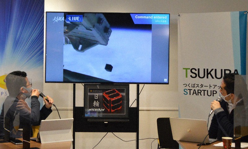 宇宙航空研究開発機構(JAXA)の配信映像から、日輪(テレビ画面下部の立方体)が宇宙空間に放出される様子を見守るワープスペース最高経営責任者の常間地悟さん(左)と、五十嵐立青・つくば市長=茨城県つくば市吾妻2で