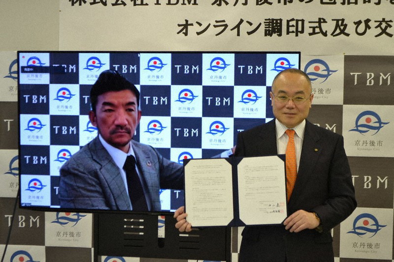 オンラインを利用し、パフォーマンスで調印書を掲げる山崎敦義CEO(左)と中山泰市長=京都府京丹後市で、松野和生撮影