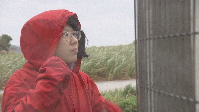 映画「ちむぐりさ 菜の花の沖縄日記」の一場面。フェンス越しに米軍基地を見つめる坂本菜の花さん=沖縄テレビ放送提供