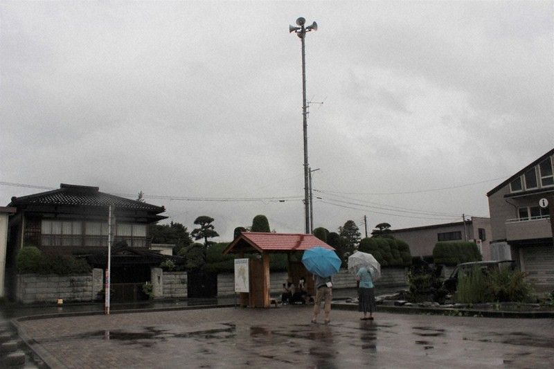 雨の中で防災行政無線から流れる「若者のすべて」のチャイムに聴き入るファン=山梨県富士吉田市の富士急行線下吉田駅前で2021年7月9日、小田切敏雄撮影