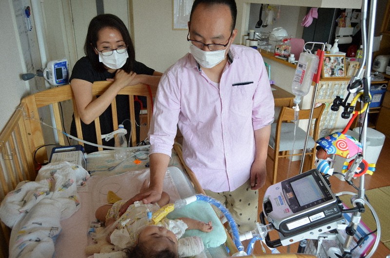 人工呼吸器を付けて自宅で過ごす陽奈子ちゃんを見守る母昌子さんと父英之さん。避難時には機器の電源確保や移動を支える人手の確保が欠かせない