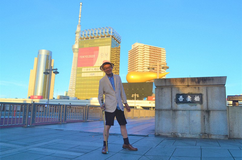 荷風も愛した隅田川にかかる「吾妻橋」でポーズをとる坂崎重盛さん。アサヒグループ本社ビルには「一歩ずつ、明日へ。」の応援メッセージが=東京・浅草で鈴木琢磨撮影