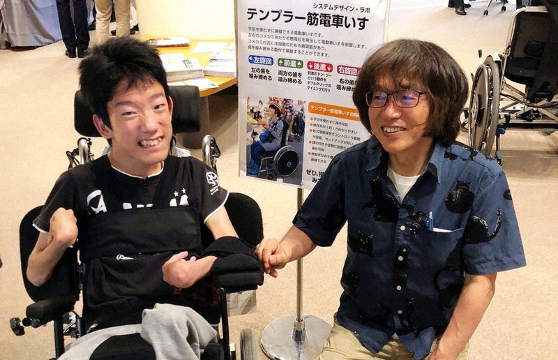 構井遼さん(左)からの要望を受け、奥歯をかみしめて操作する電動車椅子の開発に取り組んだ「魔法使い」の杉本義己さん=神戸市内で2018年5月、杉本義己さん提供
