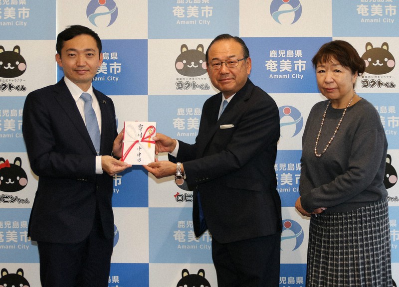 ふるさと納税100万円を安田壮平・奄美市長(左)に手渡す中津留瑞良さん(中央)と妻の直美さん=鹿児島県奄美市提供