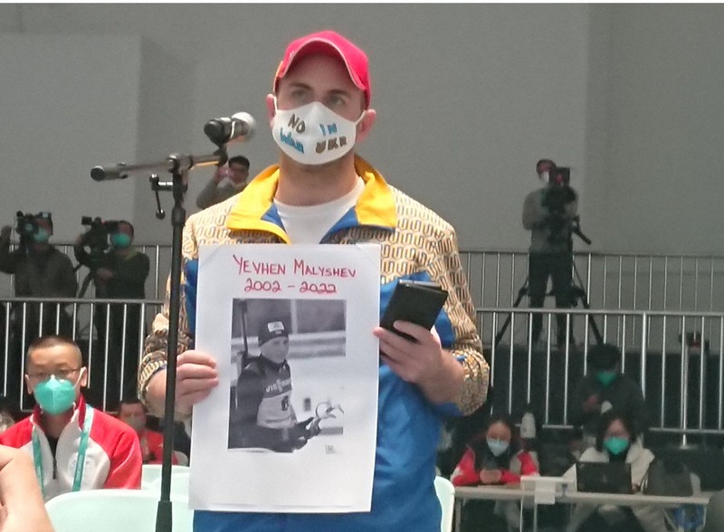 ロシアによるウクライナ侵攻で犠牲になったバイアスロン選手の写真を手に記者会見で質問するウクライナ紙の記者。マスクには「NO WAR IN UKR(ウクライナの戦争に反対)」の文字があった=北京市内で2022年3月2日午後9時40分、岩壁峻撮影