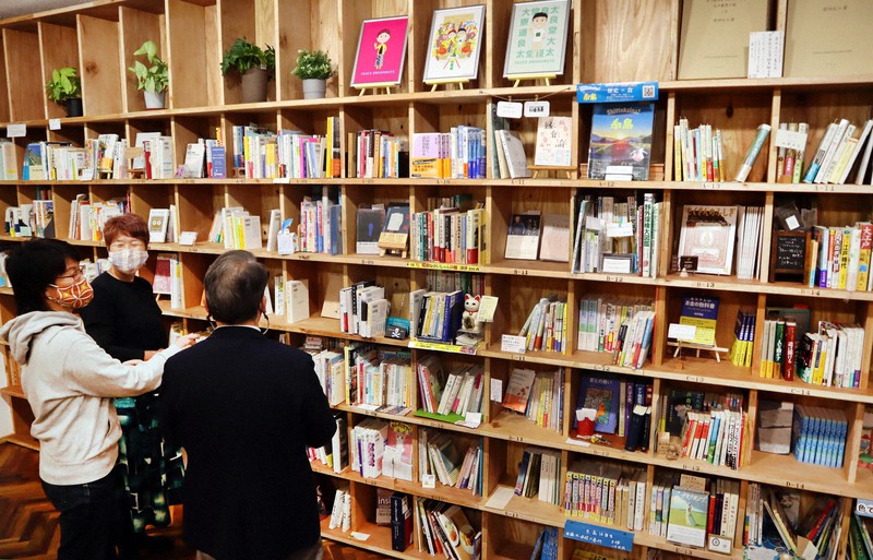 壁いっぱいに広がる書棚の前で棚のオーナーや客と談笑する「糸島の顔がみえる本屋さん」の管理人、中村真紀さん(左から2人目)。靴を脱いで上がるフローリングの床に座り、くつろぎながら本を選ぶことができる=福岡県糸島市で2021年11月22日、谷由美子撮影