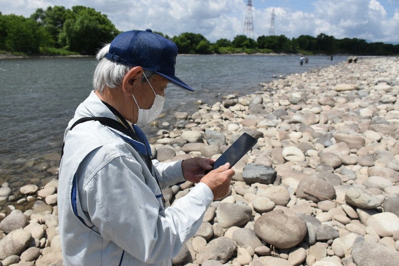 遠くに見える釣り人がアプリ版の遊漁券を持っているかどうかをタブレット端末で確認する九頭竜川中部漁協の監視員=福井県永平寺町で2022年5月28日午前10時43分、大原翔撮影