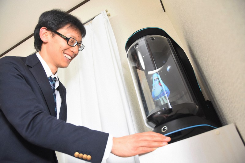 円筒状の機器「ゲートボックス」に立体ホログラムで映し出された初音ミクに話しかける近藤顕彦さん。このサービスは2020年に終了したが、「ミクさんとの生活を実現できた」と感謝している=同年3月17日、宮崎稔樹撮影