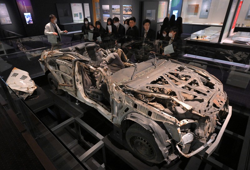 津波に襲われて殉職した佐藤雄太さんら2人の警察官が乗っていたパトカーの前で説明を聞く高校生ら＝福島県富岡町のとみおかアーカイブ・ミュージアムで2024年2月1日、手塚耕一郎撮影