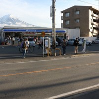 コンビニエンスストアの後ろにそびえる富士山を背景に写真撮影する外国人観光客ら＝山梨県富士河口湖町で2024年4月25日、野田樹撮影