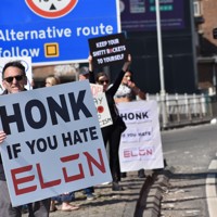 米電気自動車大手テスラの販売店前で「イーロン（・マスク氏）が嫌いならクラクションを」などと書かれたボードを車道に向けて掲げるデモ参加者＝ロンドンで2025年4月12日、福永方人撮影