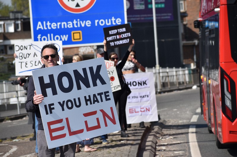 米電気自動車大手テスラの販売店前で「イーロン（・マスク氏）が嫌いならクラクションを」などと書かれたボードを車道に向けて掲げるデモ参加者＝ロンドンで2025年4月12日、福永方人撮影