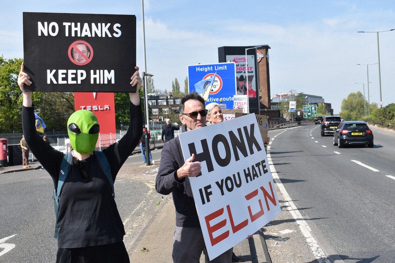米電気自動車大手テスラの販売店前で「イーロン（・マスク氏）が嫌いならクラクションを」などと書かれたボードを車道に向けて掲げるデモ参加者＝ロンドンで2025年4月12日、福永方人撮影
