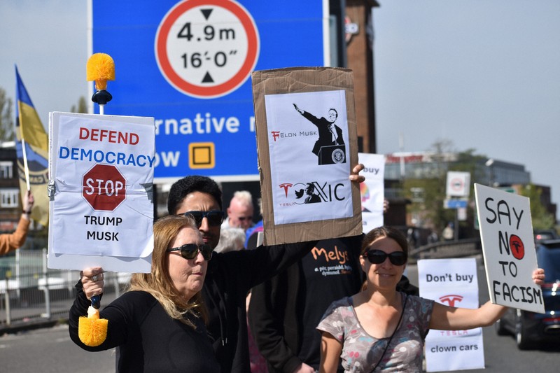 米電気自動車大手テスラの販売店前で「イーロン・マスクは有害だ」などと書かれたボードを車道に向けて掲げるデモ参加者＝ロンドンで2025年4月12日、福永方人撮影
