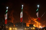 イスラエルの攻撃で石油貯蔵施設が炎上する中、揺れるイランの国旗＝テヘランで2025年6月15日、WANAロイター