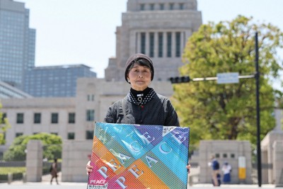 手作りのプラカードを手に国会議事堂前に立つ大木晴子さん。「スタンディング」の際は、憲法9条をイメージしたネックレスとイヤリングを必ず身につける＝東京都千代田区で2025年5月3日、幾島健太郎撮影