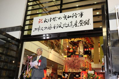 滋賀県書道協会理事長・毎日書道会評議員の神田浩山さんが筆を執ったユネスコ登録を祝う看板下であいさつする佐藤健司大津市長＝大津市中央1の大津祭曳山展示館で2025年12月11日午後6時14分、岸桂子撮影